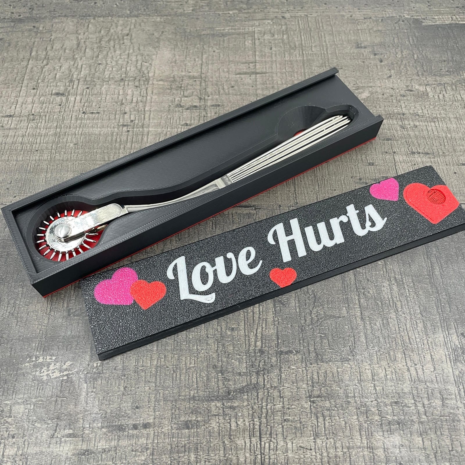 Een silverkleurige BDSM Wartenberg pinwheel in een doosje met de tekst "Love Hurts"
