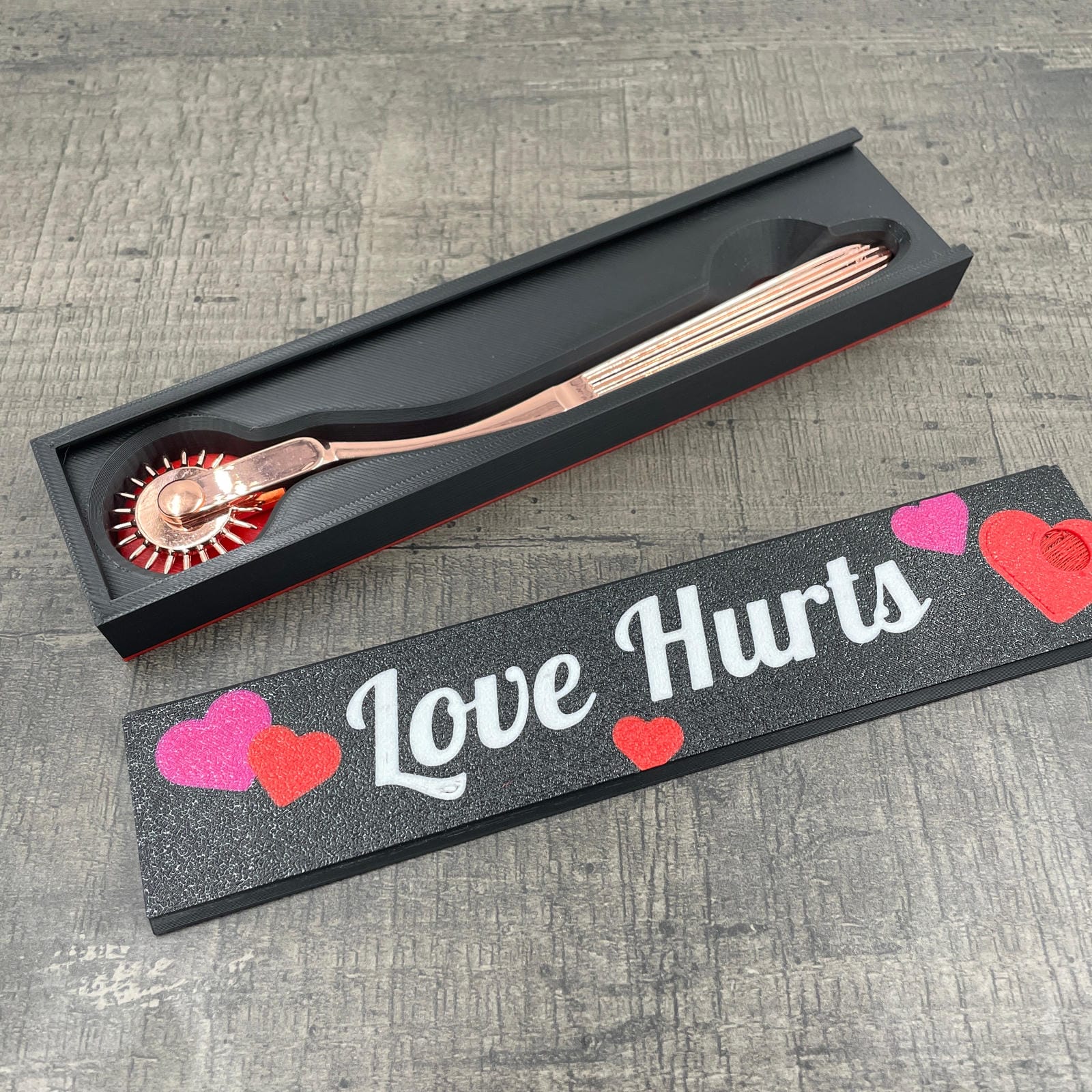 Een rose-goudkleurige BDSM Wartenberg pinwheel in een doosje met de tekst "Love Hurts"